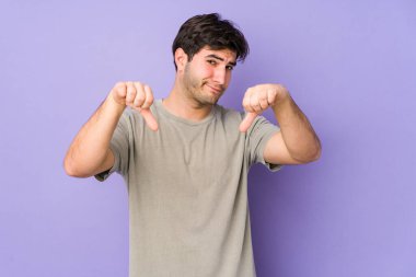Mor arka planda izole edilmiş genç adam baş parmağını aşağı gösteriyor, hayal kırıklığı konsepti..