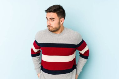 Mavi arka planda izole edilmiş genç beyaz adam omuzlarını silkeliyor ve gözleri açık..