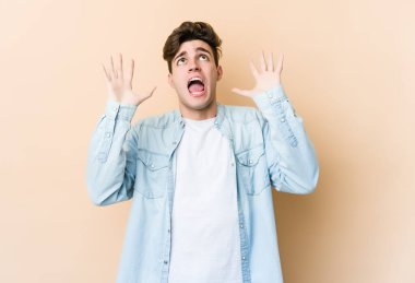 Kafkasyalı genç adam bej arka planda izole edilmiş gökyüzüne haykırıyor, yukarı bakıyor, hayal kırıklığına uğramış.