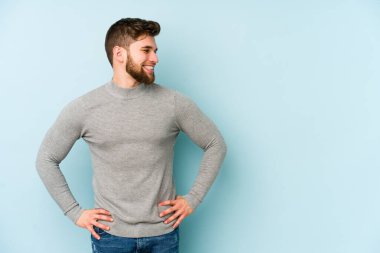 Kafkasyalı genç, mavi arka planda izole edilmiş. Kendine güvenen, kalçaları tutan biri..