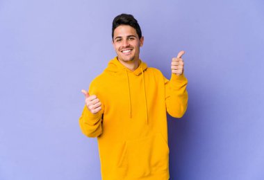 Mor arka planda izole edilmiş genç beyaz adam iki parmağını da kaldırıyor, gülümsüyor ve kendinden emin..
