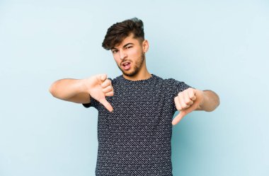 Mavi arka planda izole edilmiş genç bir Arap adam baş parmağını aşağı gösteriyor ve hoşlanmadığını ifade ediyor..