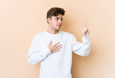 Beyaz tenli genç adam bej arka planda izole edilmiş yemin ediyor, elini göğsüne koyuyor..