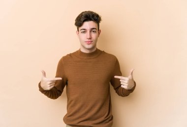 Beyazlı genç bir adam bej arka planda izole edilmiş. Elle bir gömlek fotokopi alanını işaret ediyor. Gururlu ve kendinden emin.