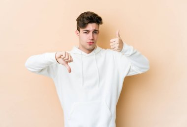 Beyaz tenli genç adam bej arka planda izole edildi. Baş parmağını kaldırdı ve baş parmağını kaldırdı. Zor seçim kavramı.