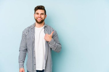 Mavi arka planda izole edilmiş genç beyaz adam gülümsüyor ve başparmağını kaldırıyor.