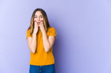 Mor arka planda izole edilmiş genç beyaz kadın şok olmuş, ağzı ellerle kaplanmış, yeni bir şey keşfetmeye hevesli..