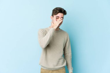 Mavi arka planda izole edilmiş genç beyaz adam kameraya parmaklarıyla göz kırpıyor, yüzünü kapatmaktan utanıyor..