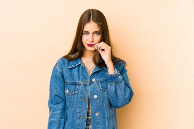Beyaz tenli genç bir kadın bej arka planda izole edilmiş ve parmakları dudaklarında bir sır saklıyor..