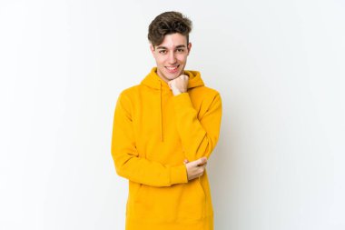 Beyaz arka planda izole olmuş genç beyaz bir adam gülümsüyor ve kendinden emin, çenesine elleriyle dokunuyor..