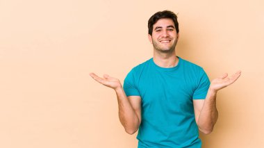 Bej arka planda izole genç adam kollarıyla ölçek yapar, mutlu ve kendinden emin hisseder..