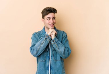 Beyaz tenli genç adam bej arka planda izole edilmiş ellerini çenesinin altında tutuyor, mutlu bir şekilde yana bakıyor..