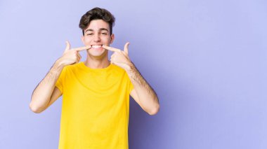 Mor arka planda izole edilmiş genç beyaz adam gülümsüyor, parmaklarını ağıza doğrultuyor..