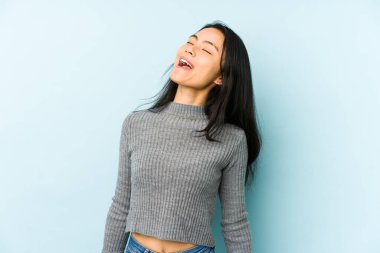 Genç Çinli bir kadın mavi bir arka planda izole edilmiş rahat ve mutlu bir şekilde gülüyor, boynunda dişlerini gösteriyor..