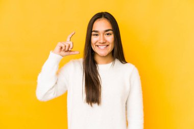 Sarı arka planda izole edilmiş genç bir kadın işaret parmaklarıyla küçük bir şey tutuyor, gülümsüyor ve kendinden emin..