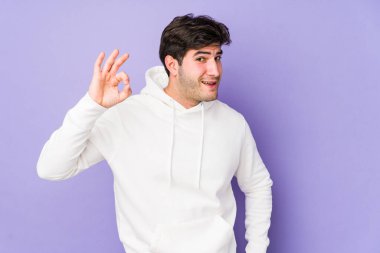 Mor arka planda izole olmuş genç bir adam bir göz kırpıyor ve elleriyle iyi bir jest yapıyor..