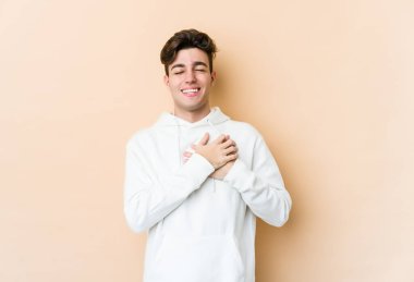 Beyazlı genç bir adam bej arka planda izole edilmiş. Gülüyor, elini kalbe koyuyor, mutluluk kavramını koruyor..