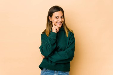 Bej arka planda izole edilmiş genç bir kadın gülümsüyor mutlu ve kendinden emin, elleriyle çenesine dokunuyor..