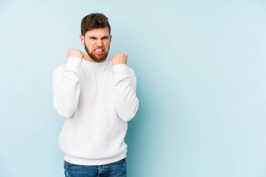 Mavi arka planda izole edilmiş genç beyaz adam yumruğunu kameraya gösteriyor, agresif yüz ifadesi var..