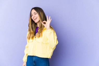 Genç beyaz kadın, mor arka planda izole edilmiş neşeli ve kendinden emin, iyi bir jest yapıyor..