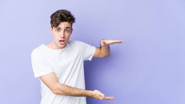 Mor arka planda izole genç beyaz adam şok ve hayret içinde elleri arasında bir kopya alanı tutuyor.