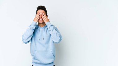 Beyaz arka planda soyutlanmış beyaz tenli genç bir adam elleriyle gözlerini kapatıyor, geniş bir gülümsemeyle bir sürpriz bekliyor..