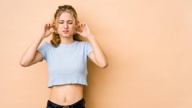 Beyaz tenli genç bir kadın bej arka planda izole edilmiş parmaklarıyla kulaklarını kapatıyor, gürültülü bir ortam yüzünden stresli ve çaresiz..