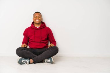 Genç latin adam yerde tek başına oturmuş kahkahalar atıyor ve gözlerini kapatıyor, rahatlamış ve mutlu hissediyor..