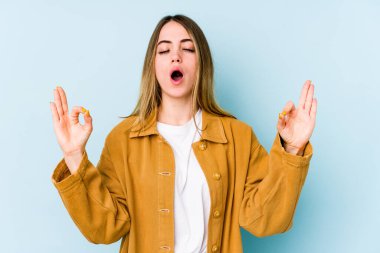 Mavi arka planda izole edilmiş genç beyaz kadın yoğun bir günün ardından rahatlıyor, yoga yapıyor..