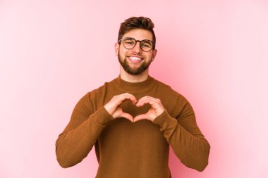 Pembe arka planda izole edilmiş genç beyaz adam gülümsüyor ve elleriyle kalp şeklini gösteriyor..