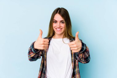 Mavi arka planda izole edilmiş genç beyaz kadın gülümsüyor ve başparmağını kaldırıyor.