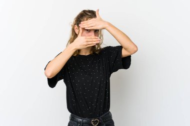 Beyaz arka plandaki genç beyaz kadın kameraya parmaklarıyla göz kırpıyor, utanarak yüzünü kapatıyor..