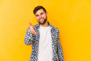 Sarı arka planda izole edilmiş genç beyaz adam selamlama jesti olarak kameraya elini uzatıyor..