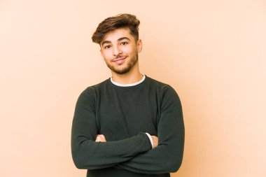 Bej bir arka planda izole edilmiş genç bir Arap, kendinden emin, kararlılıkla kollarını kavuşturuyor..