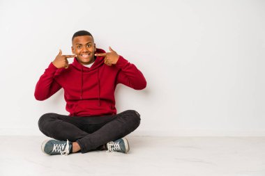 Yerde oturan genç latin adam yalnız gülümsüyor, parmaklarını ağıza doğrultuyor..