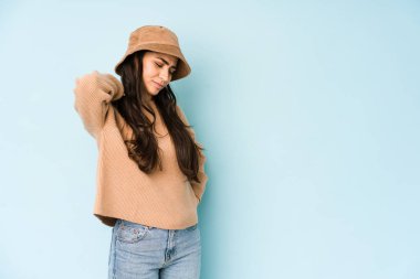 Mavi arka planda izole edilmiş bir şapka takan genç Hintli kadın stresten dolayı boynu ağrıyor, masaj yapıyor ve ona eliyle dokunuyor..