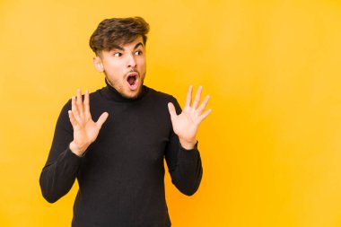 Sarı arka planda izole edilmiş genç bir Arap yüksek sesle bağırıyor, gözleri açık ve elleri gergin tutuyor..