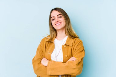Mavi arka planda izole edilmiş genç beyaz kadın kendini güvende hissediyor, kararlılıkla kollarını kavuşturuyor..