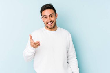 Mavi arka planda izole edilmiş genç beyaz adam selamlama jesti olarak kameraya elini uzatıyor..
