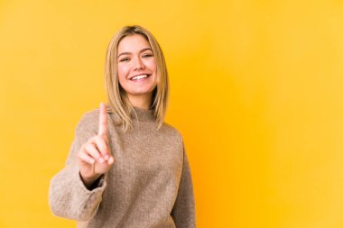 Sarışın, beyaz bir kadın, sarı arka planda izole edilmiş. Bir numarayı parmağıyla gösteriyor..