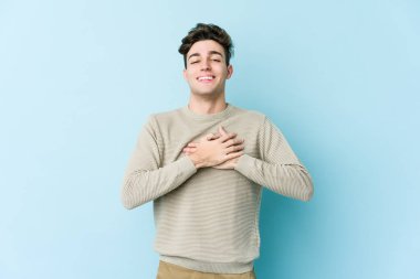 Kafkasyalı genç adam mavi arka planda izole olmuş gülüyor, elini yüreğine koyuyor, mutluluk kavramını koruyor..