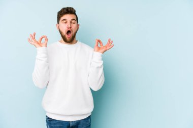 Mavi arka planda izole edilmiş genç beyaz adam yorucu bir günün ardından rahatlıyor. Yoga yapıyor..