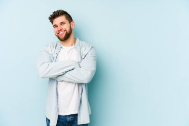 Mavi arka planda kendini güvende hisseden, kararlılıkla kollarını kavuşturan genç beyaz bir adam..