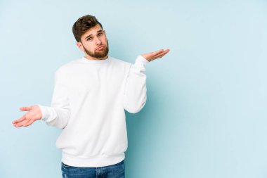Kafkasyalı genç adam mavi arka planda izole edilmiş. Kafası karışmış ve omuz silkiyor. Kopya alanını tutmak için..