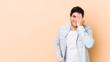 Bej arka planda izole edilmiş genç bir adam yüzünün yarısını avuç içiyle kaplayarak eğleniyor..