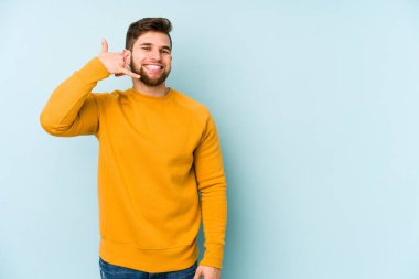 Mavi arka planda izole edilmiş genç beyaz adam parmaklarıyla bir cep telefonu hareketi gösteriyor..