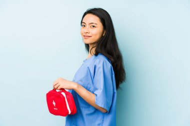 Genç Çinli hemşire mavi bir arka planda izole edilmiş şüpheli ve şüpheci bir ifadeyle yana bakıyor..