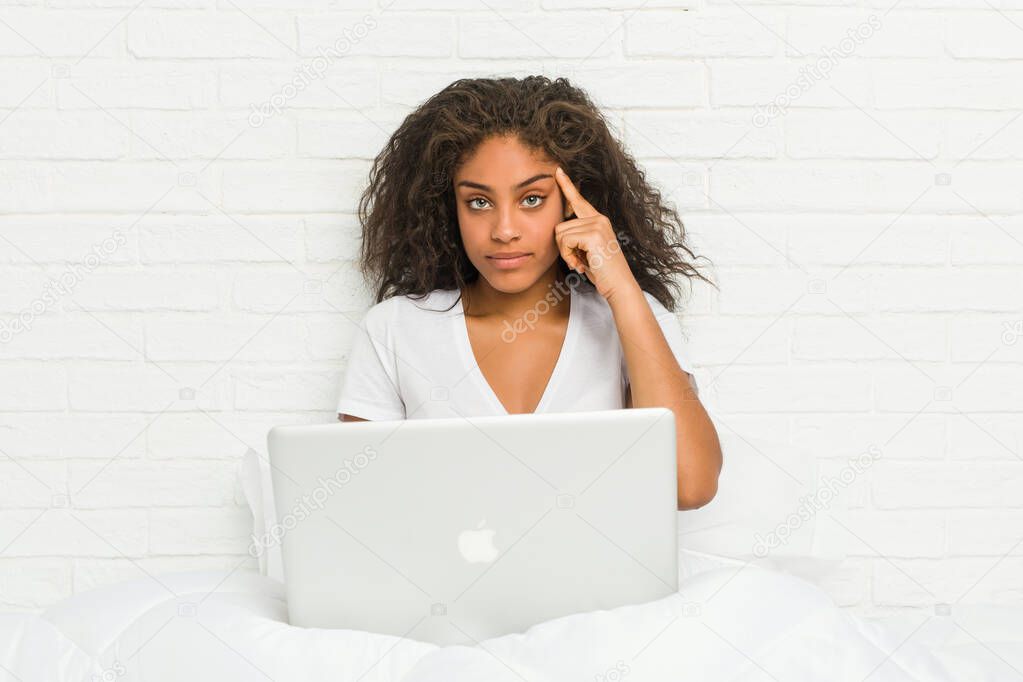 Young african american woman sitting on the bed with laptop pointing his temple with finger, thinking, focused on a task.