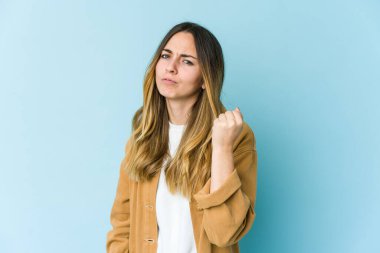 Mavi arka planda izole edilmiş genç beyaz kadın yumruğunu kameraya gösteriyor, agresif yüz ifadesi..