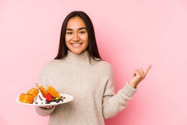 Genç Asyalı bir kadın bir waffle tutarak kendini soyutlamış gülümsüyor ve boş alanda bir şeyler gösteriyor..
